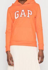 Sudadera de algodón naranja con bolsillo frontal y logo blanco de "GAP". Cuenta con una capucha con cordón y puños acanalados. Llevada sobre una falda blanca con pliegues.