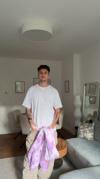 T-shirt bianco indossato da una persona che tiene un felpa tie-dye lilla. La stanza presenta un pavimento in legno chiaro, un divano grigio e elementi decorativi.