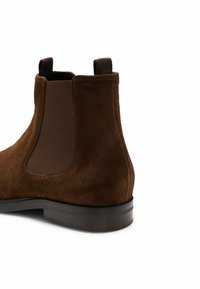 Brązowy zamszowy botek za kostkę z elastycznymi panelami bocznymi, okrągłym noskiem i ciemną gumową podeszwą. Posiada detale w postaci przeszyć na cholewce.