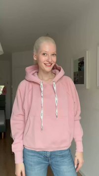 Noor naine naeratamas, kandes roosat kapuutsiga pusa valgete nöörpaeltega ja siniseid teksapükse, seistes heledas toas, kus on raamitud seinakunst.