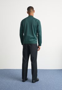 Grön, lätt jacka med ribbad krage och ärmslut, tillsammans med svarta byxor och mörka skor, sedd bakifrån mot en enfärgad bakgrund.