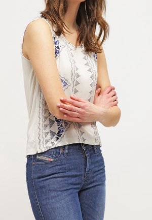 Woman in patterned sleeveless white top and blue jeans stands with arms crossed against plain background.