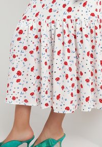 Jupe florale en tissu blanc avec des motifs de fleurs rouges et bleues, présentant un ourlet froncé et associée à des sandales à talons vertes.