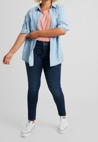 Persona arremangando las mangas de una camisa de mezclilla azul claro sobre una camiseta a rayas naranja y blanca, vistiendo jeans azul oscuro y zapatillas blancas.