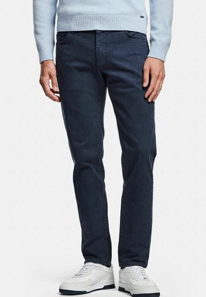 Pantalon en coton bleu foncé avec une coupe classique ; doté de cinq poches et d'une texture subtile. Associé à des baskets blanches de sport.