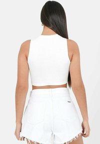 Weißes geripptes Crop-Top mit hohem Kragen, kombiniert mit weißen Jeansshorts mit fransigen Säumen und Gesäßtaschen. Schlichter Hintergrund.