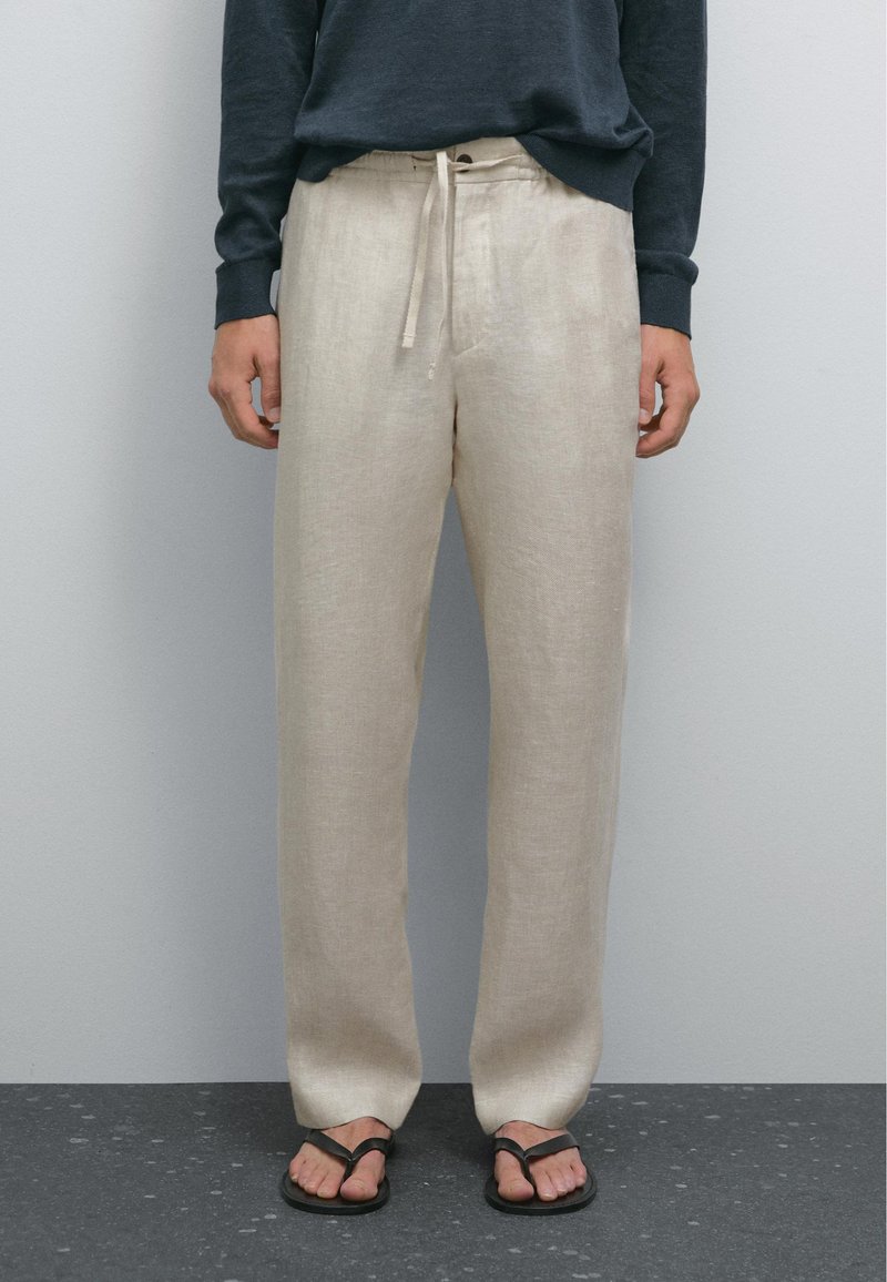 Pantalon en lin beige clair avec une coupe décontractée, ceinture à cordon de serrage et poches latérales, associé à des tongs noires. Texture minimale visible.