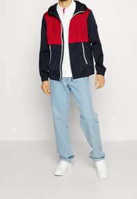 Giacca a zip color block nei toni del blu navy e rosso, con cappuccio, polsini elasticizzati e dettagli in bianco sulla chiusura. Indossata sopra una polo bianca e jeans azzurri chiari.