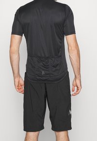 T-shirt de sport noir à manches courtes avec une poche arrière, associé à un short noir. Le tissu semble lisse et extensible.