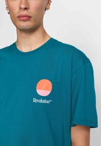 Teal katoenen t-shirt met een ronde oranje en roze grafiek op de linkerborst met de tekst "Revolutie" eronder. Eenvoudig, casual ontwerp.