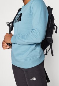 Felpa azzurra con logo nero, collo rotondo e polsini a coste. Zaino nero con spallacci regolabili indossato sulla schiena. Pantaloni neri con logo.