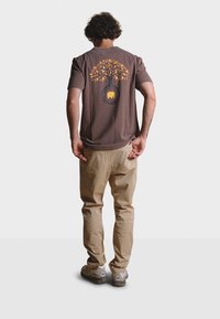 Hnědé bavlněné tričko se vzorem stromu se žlutými listy a siluetou slona, doplněné o béžové kalhoty a šedé turistické boty.