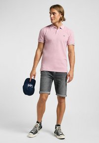 Polo shirt rose clair avec des bordures blanches, associé à un short en denim gris. Tient une casquette marine avec un texte blanc. Porte des baskets montantes noires.