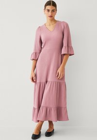 Roze, gestructureerde midi-jurk met een V-hals en 3/4 mouwen in belvorm. Beschikt over een tiered rucheszoom en een relaxte pasvorm. Gecombineerd met zwarte platte schoenen.