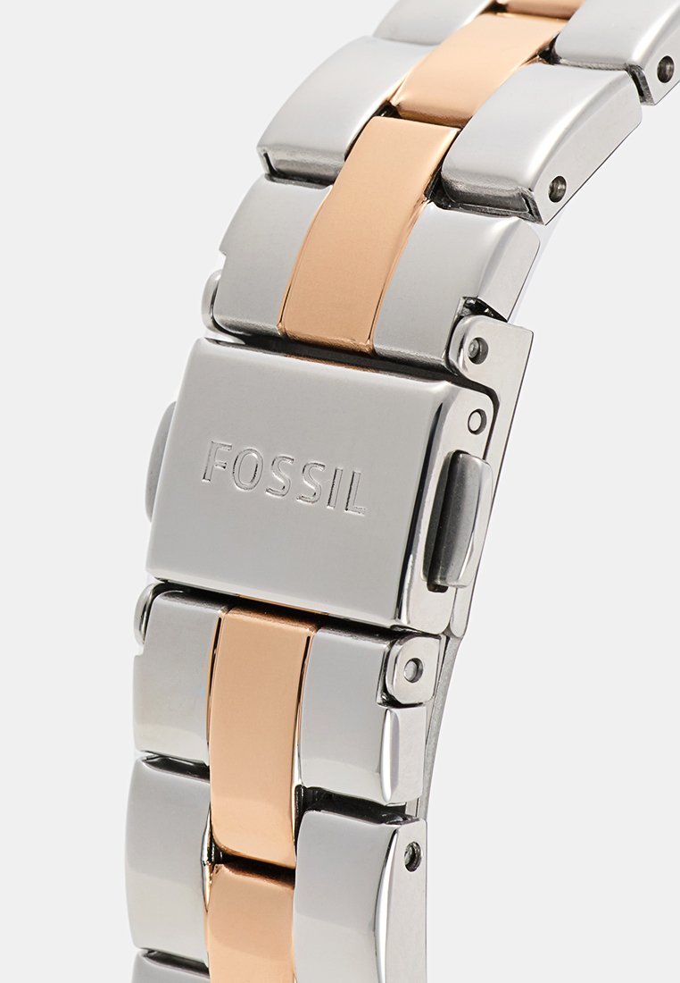 Bracelet de montre en métal argenté et or rose avec des maillons polis, arborant le logo gravé "FOSSIL" et un mécanisme de fermoir pour un ajustement sécurisé.