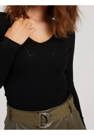 Kobieta w czarnym prążkowanym swetrze z dekoltem w serek i oliwkowozielonym pasku z podwójną srebrną klamrą w kształcie litery D, ręka przy dekolcie.