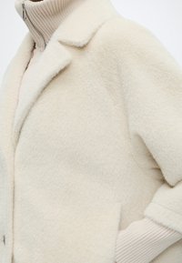 Manteau duveteux couleur crème porté par-dessus un pull à col roulé côtelé beige avec une fermeture éclair visible au niveau du cou.
