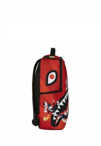 Mochila roja con acentos negros, que presenta personajes de dibujos animados y un gran gráfico de boca estilizada. El material parece suave con correas acolchadas.