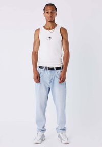 Canotta bianca con logo, jeans in denim azzurro chiaro, cintura nera e sneakers grigie. Il modello è in piedi davanti a uno sfondo bianco.