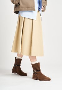 Botines de ante marrones con cremallera lateral, acento de cinta negra y tacón de madera bajo. Combinados con una falda beige y calcetines color crema.