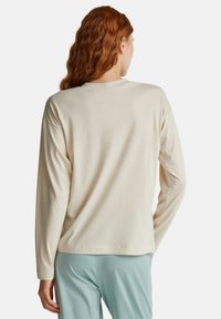 Osoba o długich, czerwonych włosach nosi luźną, kremową bluzkę z długim rękawem i jasnoniebieskie spodnie, przedstawiona z tyłu na jednolitym tle.