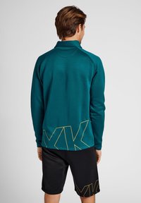 Mouwloze teal sportshirt met lange mouwen en een geel geometrisch patroon op de achterkant, gecombineerd met zwarte shorts met logo-detail. Glad textuur van de stof.