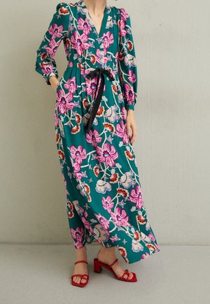 Vrouw draagt groene maxi-jurk met grote roze bloemenprint, zwarte tailleband en rode sandalen met hak, staand tegen een effen muur.