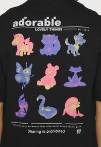 Musta T-paita, jossa on eloisia, värikkäitä kuvastoja erilaisista eläimistä ja leluista, tekstinä "suloinen" ja "Jakaminen on kielletty."
