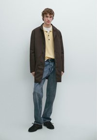 Brauner Baumwoll-Trenchcoat über einem gelben Poloshirt, kombiniert mit hellblauen Jeans und dunklen Schuhen. Glatte Textur, lässige Passform.