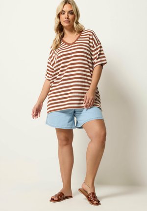 Kvinde med blondt hår iført en brun- og hvidstribet løs top, lyseblå denimbukser og brune sandaler, stående mod en ensfarvet baggrund.