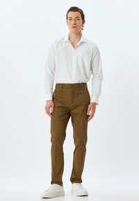 Hvid langærmet skjorte med en lomme, parret med brune tapered bukser og hvide sneakers. Enkel design, glat tekstur, struktureret pasform.