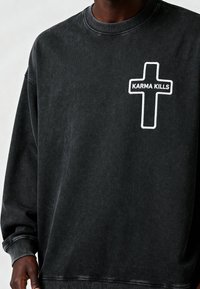 Sweat-shirt gris foncé avec une croix en contour blanc et le texte "KARMA KILLS" sur le côté gauche de la poitrine, porté par une personne à la peau foncée.