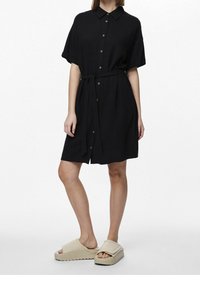 Abito camicia nero con maniche corte, vita cintrata e gonna a ruota. Indossato con sandali beige con suola texturizzata.