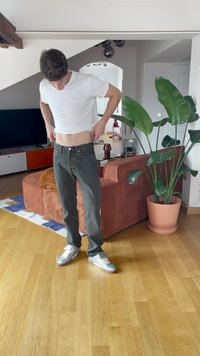 Homem de camiseta branca e jeans escuros a ajustar as calças numa sala de estar com um sofá castanho e uma grande planta verde em vaso.