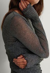 Femme portant un haut noir et argenté pailleté ajusté à manches longues avec des manches transparentes, tenant son cou d'une main et sa taille de l'autre.