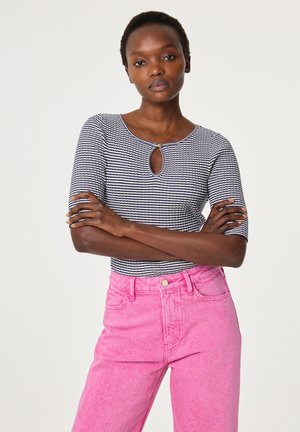 Vrouw met kort haar staat met gekruiste armen, draagt een zwart-wit gestreepte top met een sleutelgatdetail en een hoge taille roze spijkerbroek.