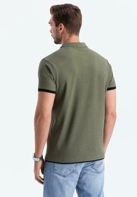 Polo shirt verde oliva con dettagli neri nel colletto, maniche corte e una trama solida, abbinato a jeans in denim azzurro chiaro.
