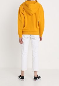 Osoba stojąca tyłem, ubrana w musztardową bluzę z kapturem, białe krótkie dżinsy i czarne buty sportowe niskie z białymi podeszwami.