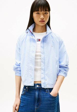 Lichtblauwe nylon jas met een staande kraag, elastische manchetten en drukknoopsluitingen. Gekörd over een wit t-shirt met een rood en blauw logo, gecombineerd met denim.