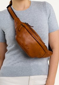 Brun läder crossbody-väska med en dragkedjeficka, rundad form och justerbar rem, bärs över en grå tröja.