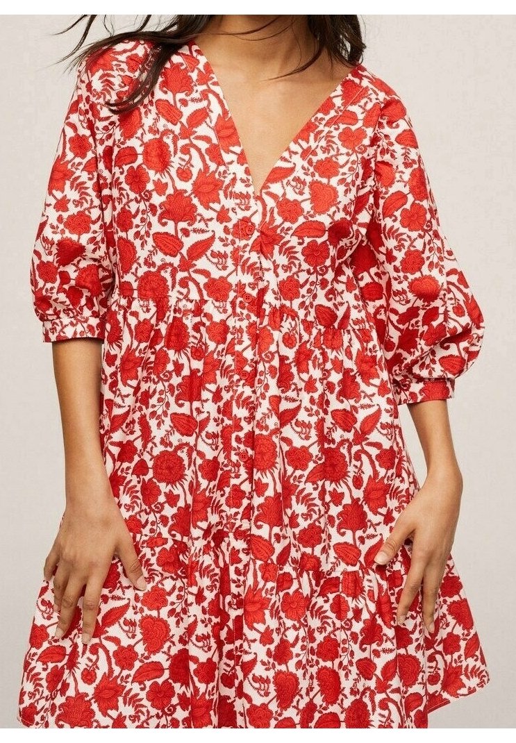 Femme portant une robe blanche avec des motifs floraux rouges vifs et des manches bouffantes trois-quarts, debout avec les mains détendues le long du corps.