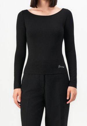 Frau trägt ein eng anliegendes schwarzes Langarmshirt mit weitem Ausschnitt und schwarze Hosen, mit "Guess" auf der unteren Seite bestickt.
