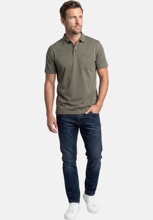 Uomo che indossa una polo verde oliva, jeans blu scuro e scarpe sportive bianche, in piedi con una mano in tasca e guarda verso sinistra.