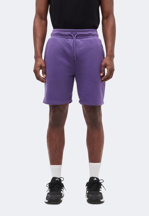 Muškarac obučen u ljubičaste šorceve sa špagom, crnu majicu bez rukava, bijele čarape i crne Adidas tenisice, stoji ispred jednobojne pozadine.