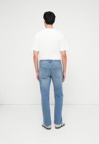 Άνδρας που στέκεται με την πλάτη προς τα εμάς, φοράει λευκό πουκάμισο με κοντά μανίκια, τζιν ανοιχτό μπλε κ αι ανοιχτόχρωμα σανδάλια με κάλτσες, μπροστά σε ένα απλό φόντο.