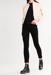 Veste bomber beige clair avec fermeture éclair, jeans noirs ajustés et bottines en cuir noir à la cheville avec une semelle de couleur naturelle.