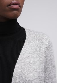 Pletený šedý kardigan s jemnou texturou, nošený přes černý rolák. Má výstřih do V a volný střih. Minimalistický styl.