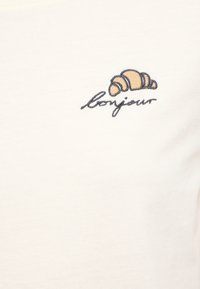 White cotton shirt featuring a brown embroidered croissant and the word "bonjour" in a cursive font beneath it.