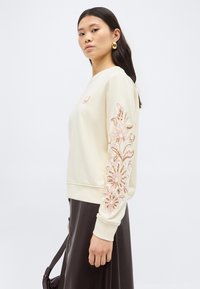 Γυναίκα με κρεμ φούτερ με ροζ κεντήματα με λουλούδια στο μανίκι και σκουρόχρωμη φούστα, κρατώντας μια σκοτεινή τσάντα στο πλάι της.