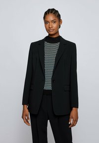 Black blazer with notched lapels, single button closure, and two front pockets, paired with a patterned knit top featuring blue and beige accents.
